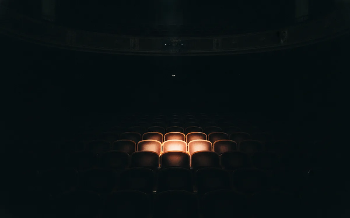 Rzędy pustych, ciemnych foteli w teatrze; jeden fotel oświetlony ciepłym pomarańczowym reflektorem